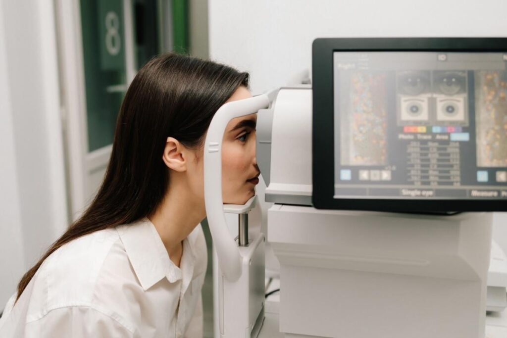 A person sits at an eye examination machine in a clinic, resting their chin on a support while looking into the device as a screen displays eye scan data.